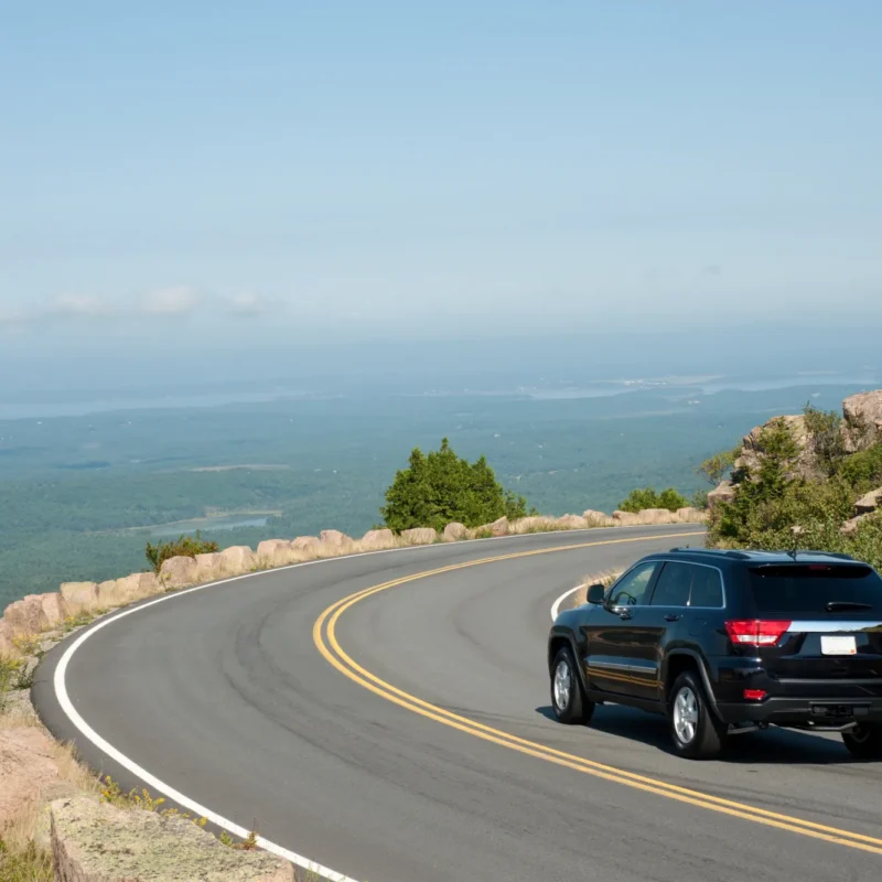 A black SUV driving around a curve on a two-lane mountain road above a forested valley and distant coastline on a clear summer day.