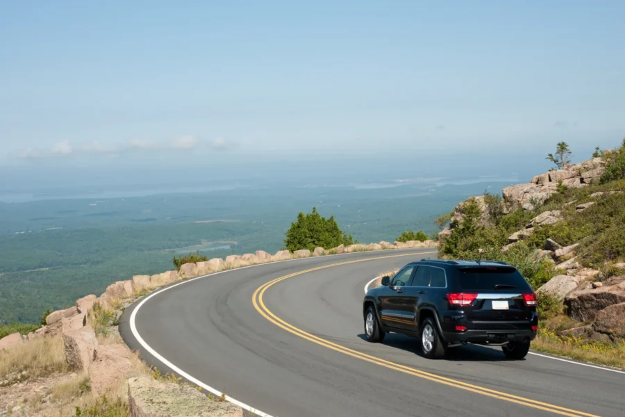 A black SUV driving around a curve on a two-lane mountain road above a forested valley and distant coastline on a clear summer day.