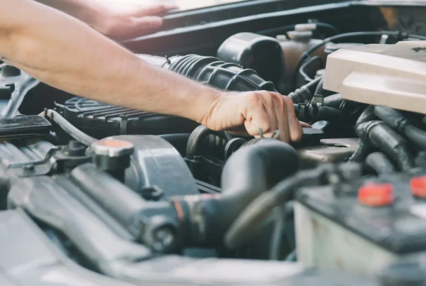 A mechanic's arm and hand reaching into a car's open engine bay among hoses and the air filter housing.