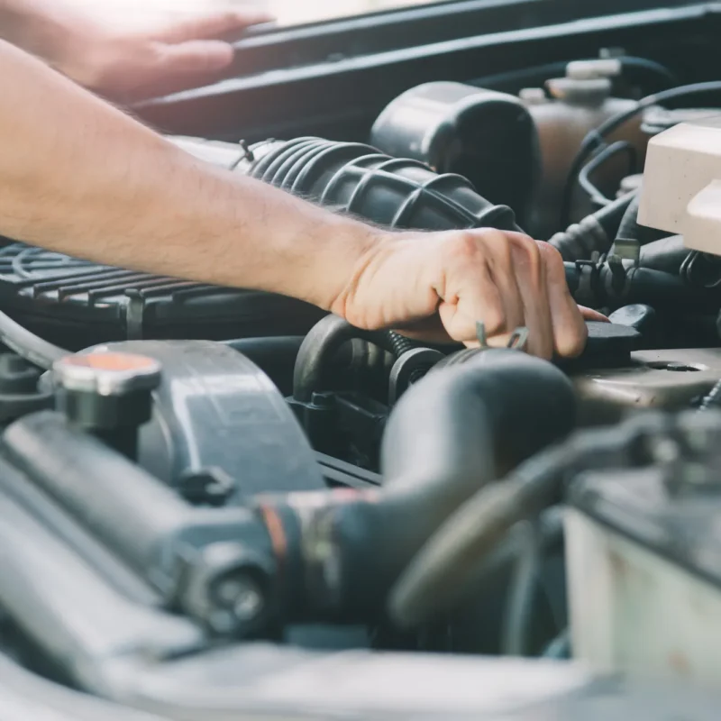 A mechanic's arm and hand reaching into a car's open engine bay among hoses and the air filter housing.