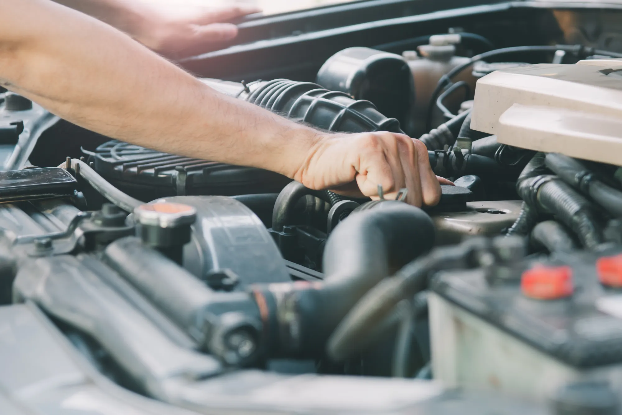 A mechanic's arm and hand reaching into a car's open engine bay among hoses and the air filter housing.