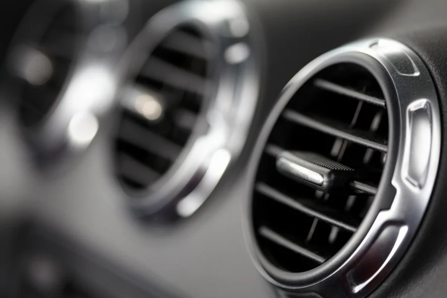 Close-up of a round chrome-trimmed air conditioning vent on a black car dashboard, with a second vent blurred in the background.