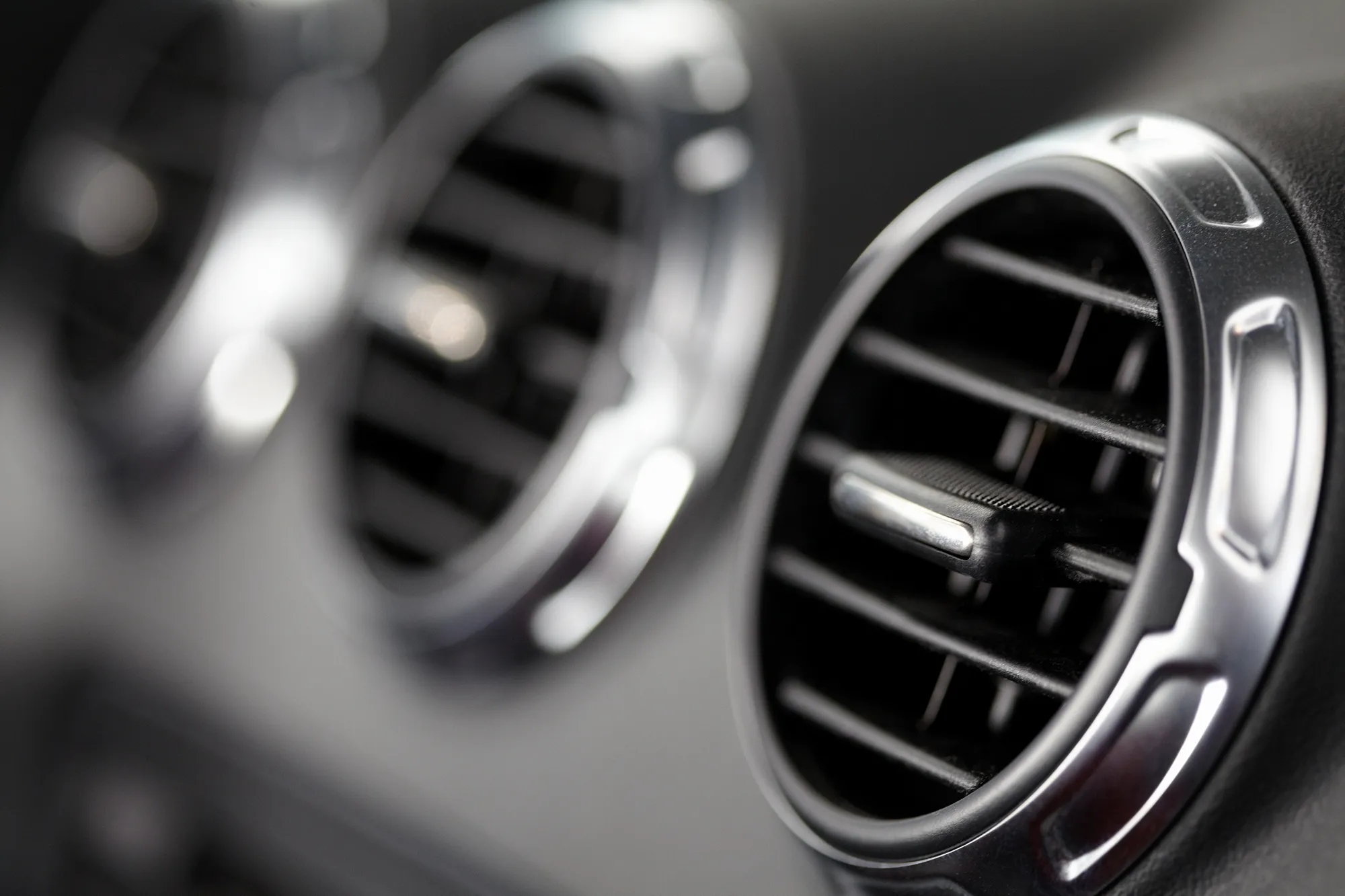 Close-up of a round chrome-trimmed air conditioning vent on a black car dashboard, with a second vent blurred in the background.