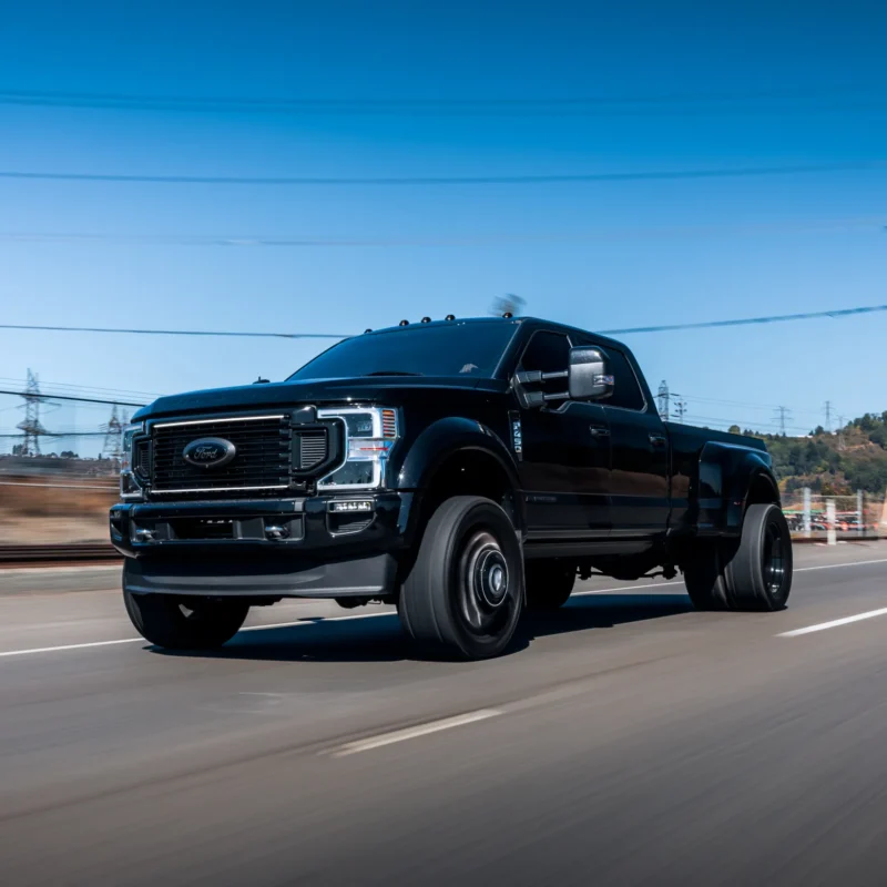 A black Ford F-450 dually pickup truck driving down a road on a clear sunny day with the background blurred by motion.