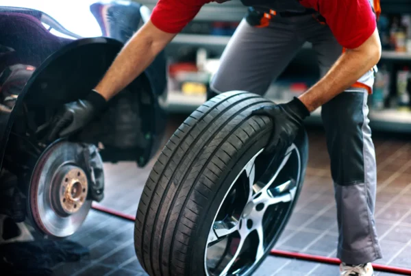 A mechanic wearing black gloves rolls a new tire on an alloy wheel toward a car with its wheel removed, exposing the brake rotor and hub, inside a repair shop.