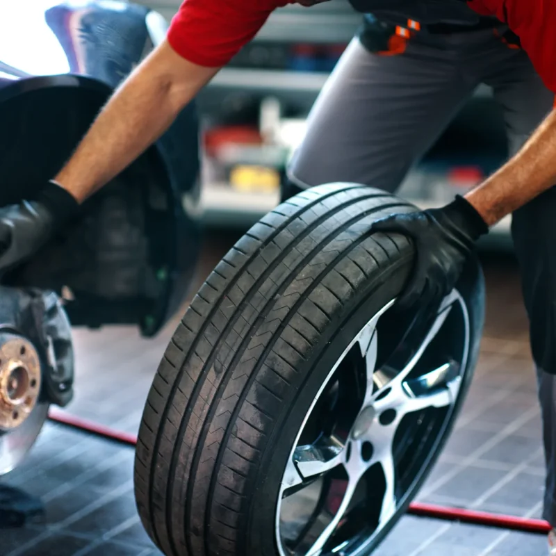 A mechanic wearing black gloves rolls a new tire on an alloy wheel toward a car with its wheel removed, exposing the brake rotor and hub, inside a repair shop.