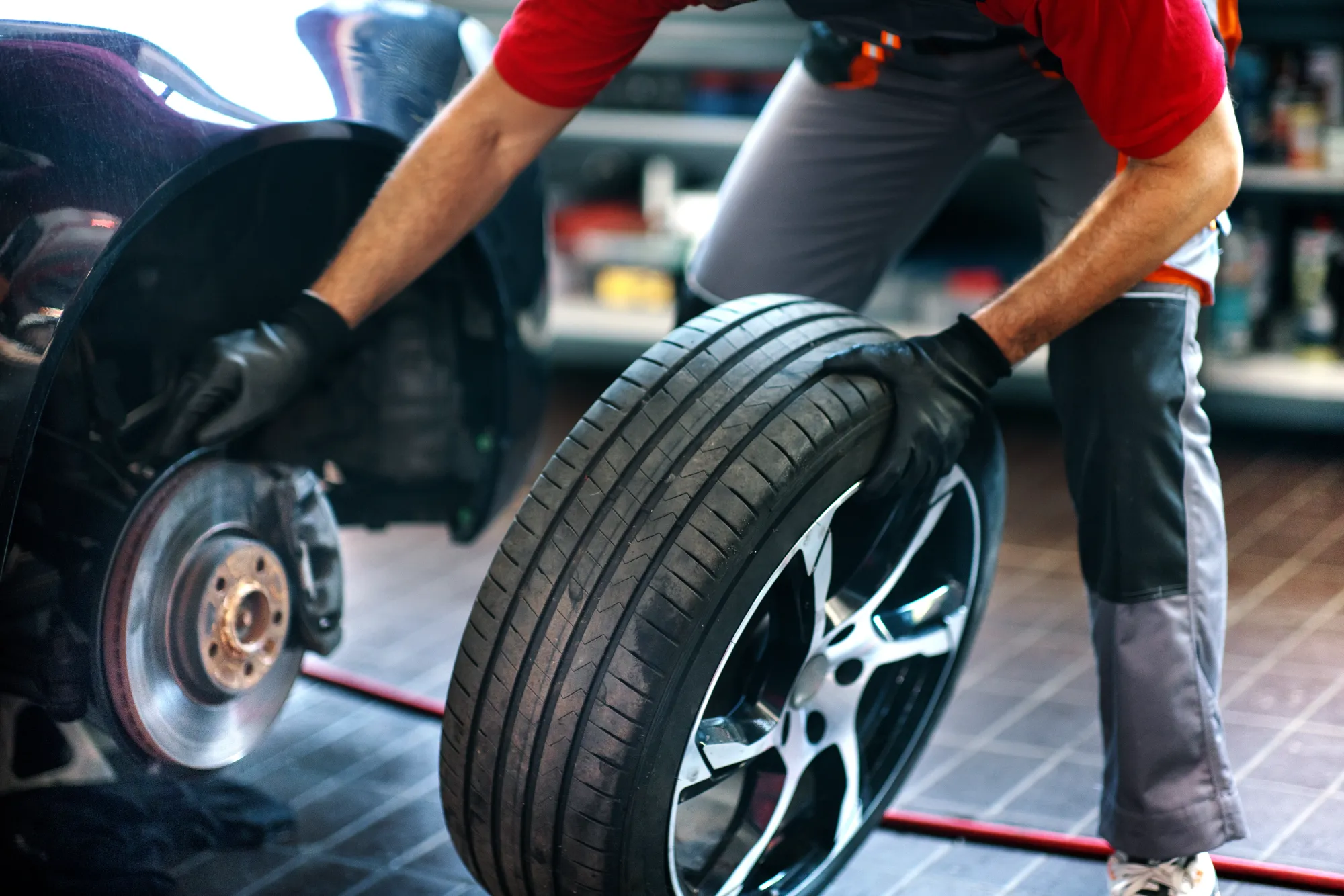 A mechanic wearing black gloves rolls a new tire on an alloy wheel toward a car with its wheel removed, exposing the brake rotor and hub, inside a repair shop.