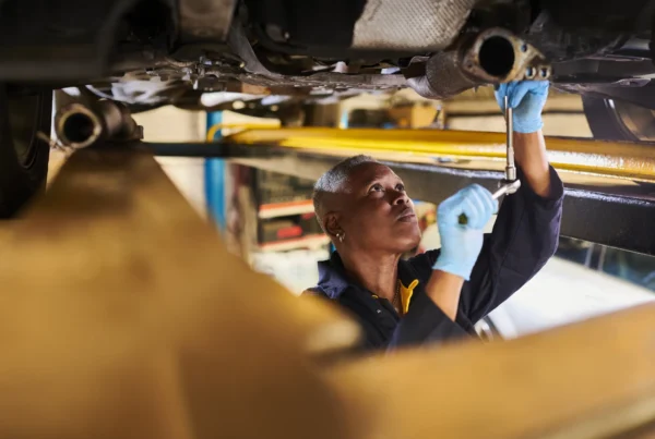 A mechanic in blue gloves uses a socket wrench on the exhaust pipe under a car raised on a shop lift.