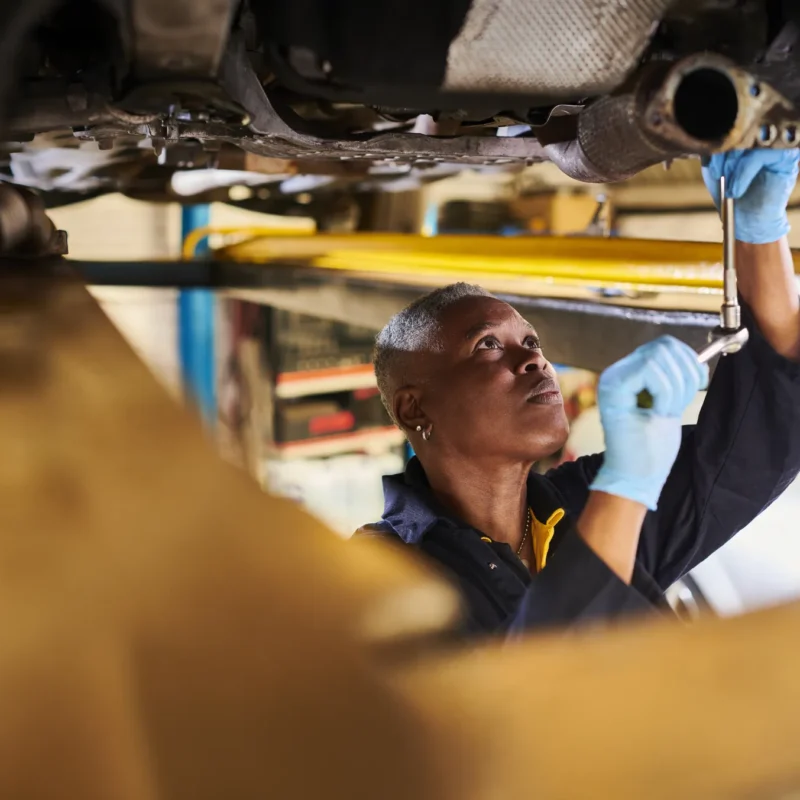 A mechanic in blue gloves uses a socket wrench on the exhaust pipe under a car raised on a shop lift.
