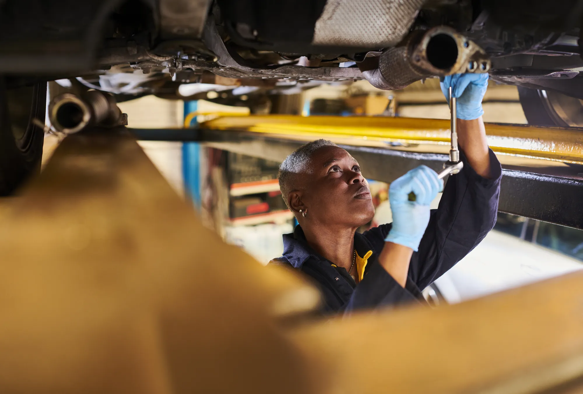 A mechanic in blue gloves uses a socket wrench on the exhaust pipe under a car raised on a shop lift.