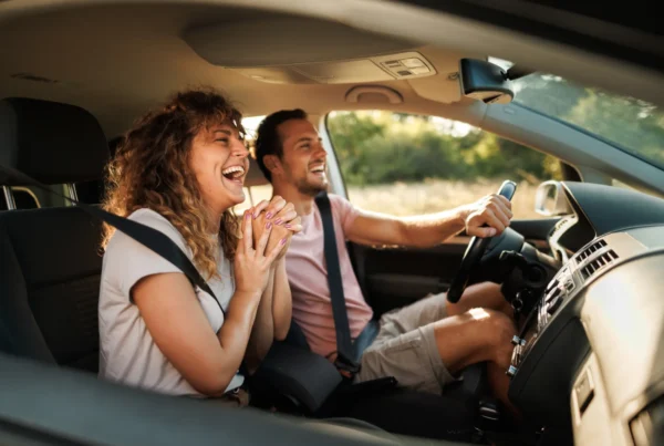 A smiling man drives a car while a laughing woman sits in the passenger seat, both wearing seat belts with green scenery outside the windows.