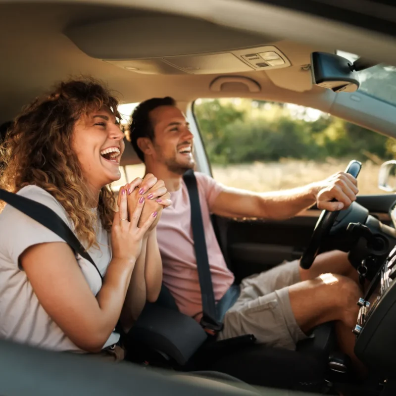 A smiling man drives a car while a laughing woman sits in the passenger seat, both wearing seat belts with green scenery outside the windows.