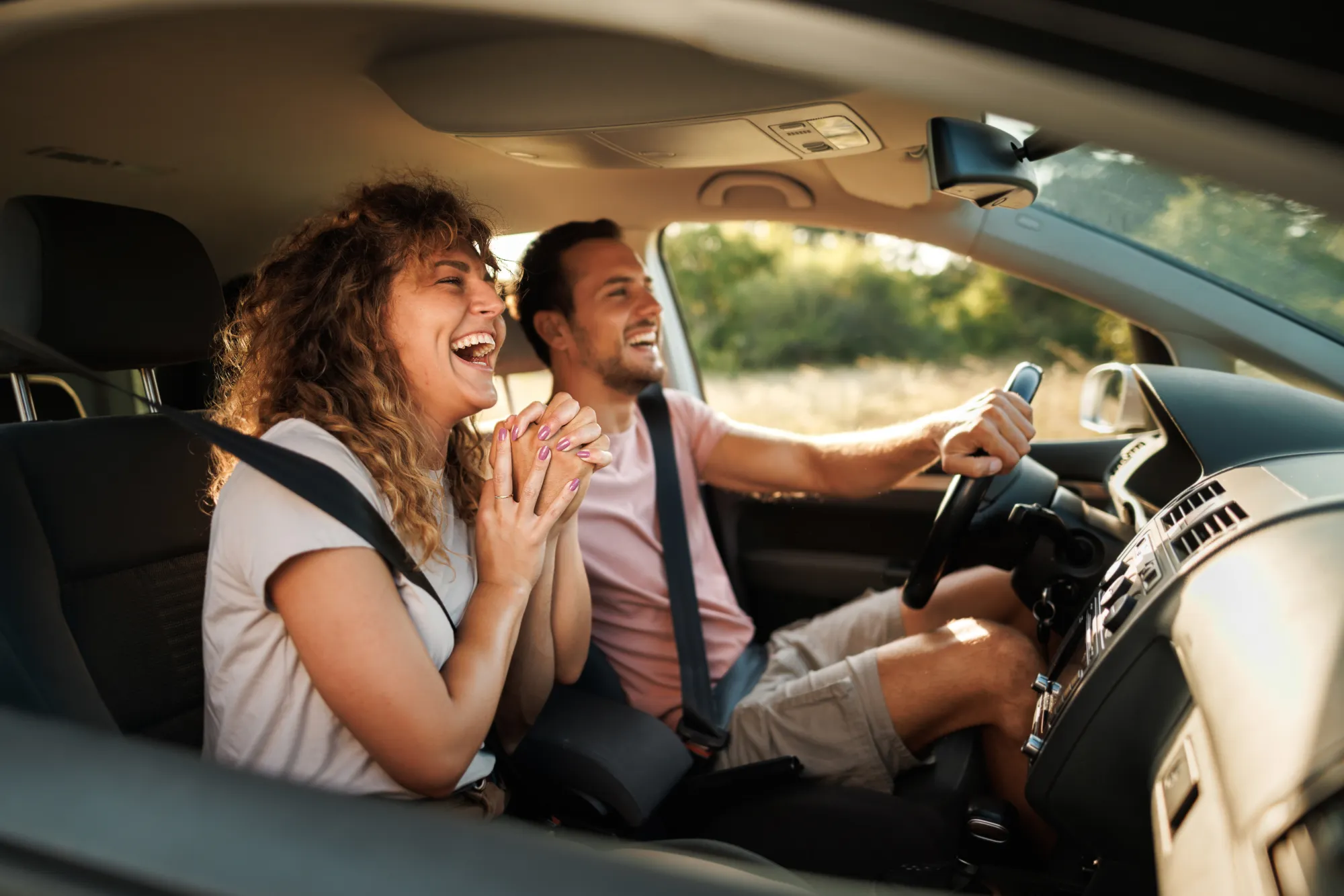 A smiling man drives a car while a laughing woman sits in the passenger seat, both wearing seat belts with green scenery outside the windows.